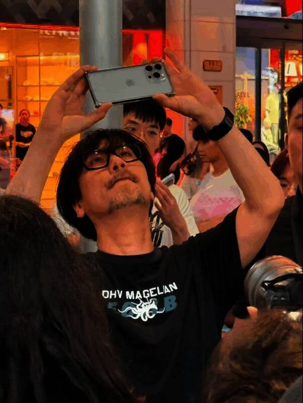 小岛秀夫在上海街头手持相机拍摄，身着黑色服装，神情专注