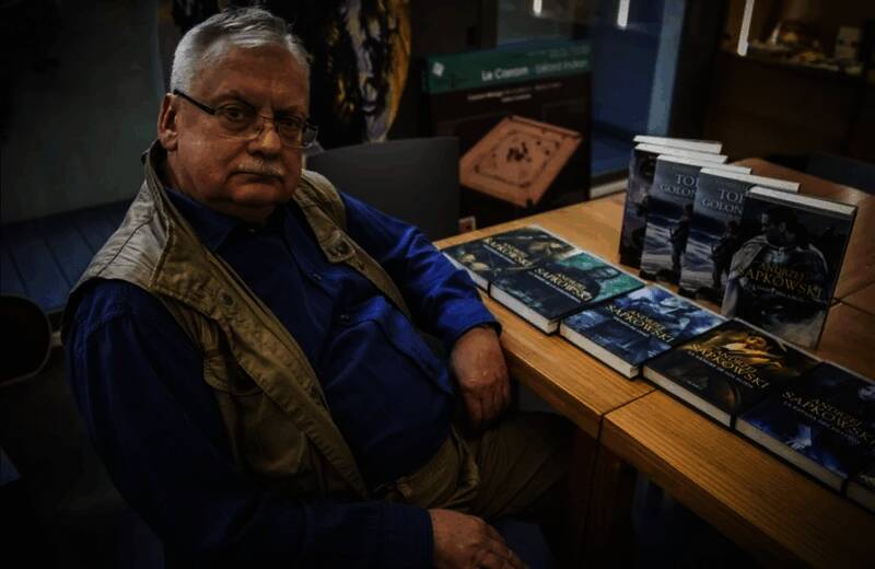 安杰伊·萨普科夫斯基正在接受采访的画面，身后是《巫师》系列书籍展示