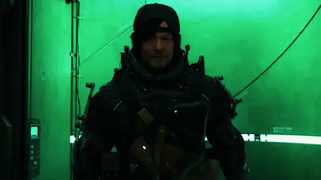 小岛秀夫站在《死亡搁浅2》展台前，身旁摆放着初代游戏的纪念品