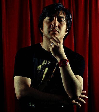 须田刚一微笑面对镜头，背后是游戏开发界面与概念图