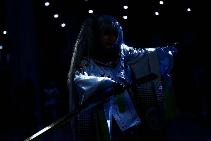 coser猫九酱扮演莉芙，身穿蓝色机械风战斗服，手持能量武器，背景为展台LED屏幕播放的游戏画面