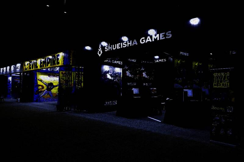 展区内玩家围站在试玩台前专注体验游戏，背景是各参展公司的品牌标识和宣传海报