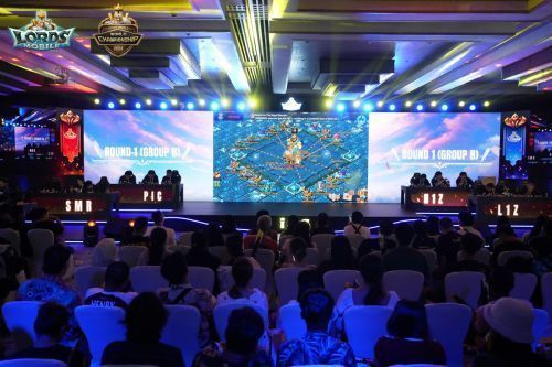 选手在比赛区专注操作手机，屏幕显示《王国纪元》实时战斗画面，周围有裁判与工作人员巡视