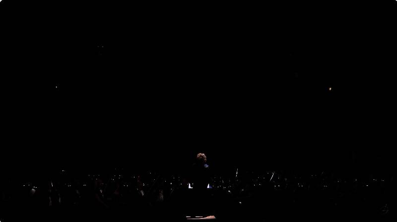 波西米亚互动25周年音乐会现场，交响乐团正在演奏游戏音乐，背景大屏幕上闪过《武装突袭4》的LOGO