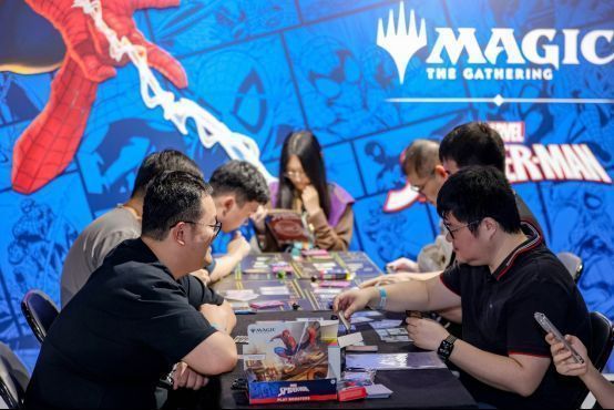 观众在展览互动区体验万智牌对战，工作人员在一旁进行规则讲解，桌上摆放着卡牌和教学手册