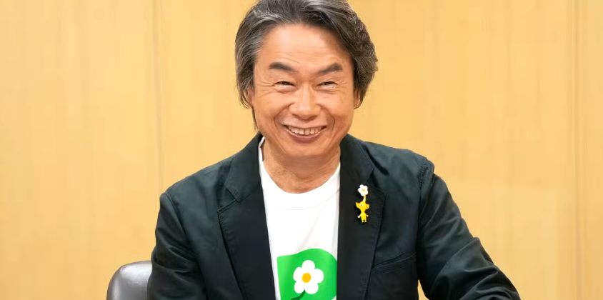 宫本茂在《Fami通》专访现场照片，身着标志性格子衬衫，面带温和微笑，背景可见《超级马里奥》经典砖块墙与Q版蘑菇图标